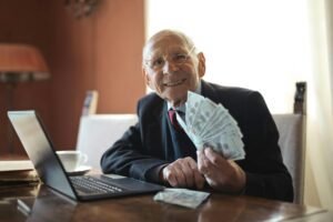 Happy senior businessman holding money in hand while working on laptop at table